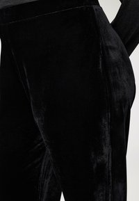 Zbliżenie na osobę w dopasowanych czarnych, welurowych spodniach, podkreślające teksturę materiału i detale szwów na neutralnym tle.