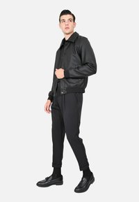 Giubbotto bomber in pelle nera con polsini e orlo a costine, indossato sopra pantaloni e scarpe nere, su uno sfondo bianco.