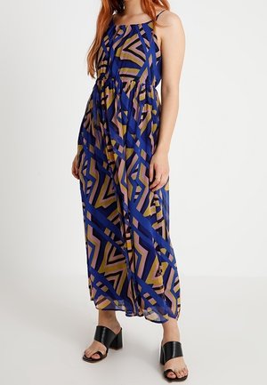 Kobieta ubrana w bezrękawnikową, długą sukienkę z geometrycznym wzorem w odcieniach niebieskiego, różowego i oliwkowego, w czarnych sandałach na obcasie, stojąca na białym tle.