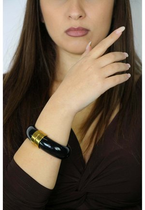 Bracelet manchette noir à finition brillante avec des accents dorés, porté au poignet. Présente une texture lisse et une forme arrondie.