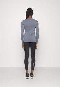 Haut gris à manches longues associé à des leggings noirs. Les leggings présentent un logo blanc sur la jambe inférieure. Le matériau semble extensible.