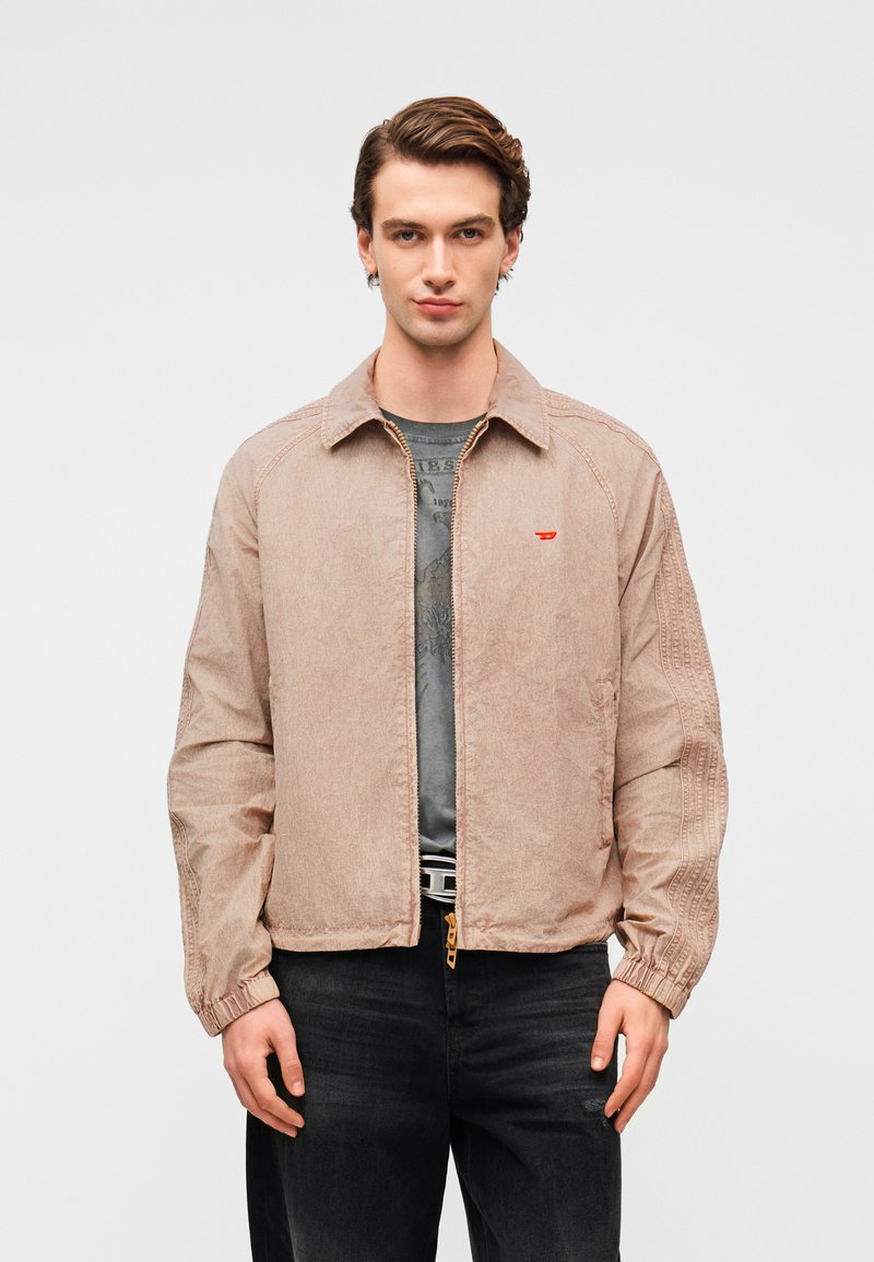 Μπεζ ελαφρύ μπουφάν με γιακά, ελαστικά μανσέτα και μικρό κόκκινο λογότυπο. Φορεμένο πάνω από γκρι μπλουζάκι και συνδυασμένο με σκούρο τζιν παντελόνι.