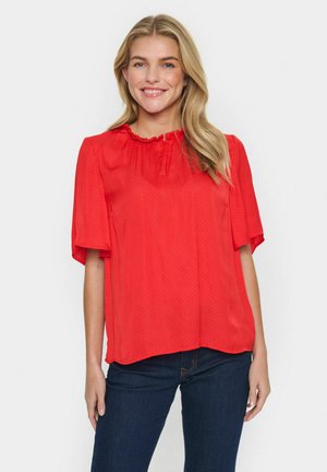 Blouse rouge avec un col volanté et des manches courtes. Fabriquée en tissu lisse avec un motif à pois. Associée à un jean bleu foncé.