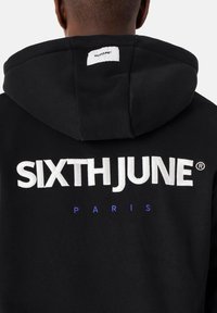 Sudadera negra con un gran logotipo bordado en blanco "SIXTHJUNE" y "PARIS" en azul debajo. Cuenta con una capucha y una textura suave.