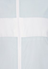 Jacket de culoare albastră deschisă, cu o dungă albă orizontală, dintr-un material texturat și cu un închizător central cu fermoar. Finisaj neted cu detalii de cusătură ordonată.