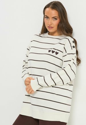 Donna con maglia bianca a maniche lunghe con strisce orizzontali nere, tre piccoli cuori neri sul petto e bottoni neri sulla spalla, in posa contro una parete bianca.