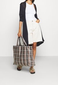 Stor brun rutig tygväska med vita detaljer, tillverkad i tyg. Har dubbla handtag. Modellen bär vit linne och ljusa shorts.