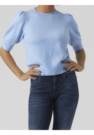 Vrouw draagt een lichtblauwe trui met korte mouwen en geplooide schouders en donkerblauwe jeans, staande met handen in de zij tegen een effen achtergrond.
