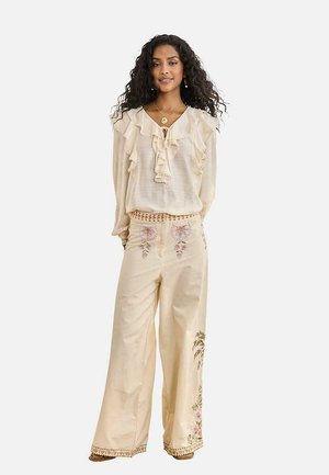 Mujer con cabello rizado, blusa crema con volantes y pantalones de pierna ancha con bordado floral, posando frente a un fondo blanco.