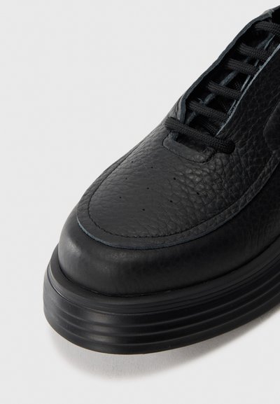 Black leather shoe with thick sole, round toe, stitched detailing, and black laces on a white background.
