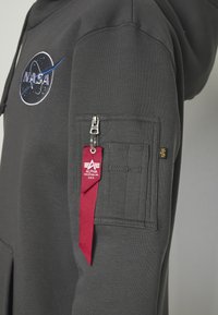 Sudadera gris con el logo de la NASA bordado en azul y blanco. Presenta un bolsillo con cremallera en la manga, cinta de acento roja y una tela estructurada.