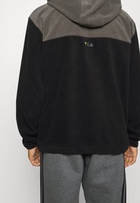 Sort og grå hættetrøje med en blød tekstur. Har et broderet logo på ryggen og en afslappet pasform, parret med grå sweatpants.