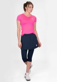 Top deportivo de mangas cortas en un brillante color rosa, falda azul oscuro y leggings capri en navy. Presenta un logo en la falda. Zapatillas deportivas completan el conjunto.