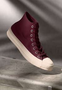 Baskets montantes en toile bordeaux avec une finition texturée, embout en caoutchouc crème, et œillets en métal pour les lacets, présentées sur un fond minimal.