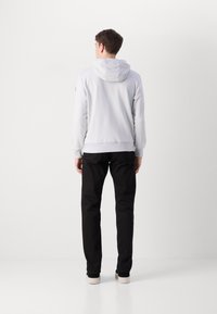 Hoodie cinza claro com capuz ajustável, punhos e bainha canelados, combinado com calças pretas e sapatos claros, visto de costas.
