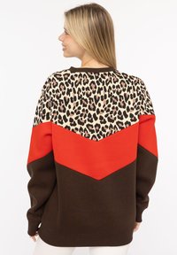 Bluza z nadrukiem w panterkę na górze, z czerwoną wstążką i brązową dolną częścią. Dzianinowa tekstura, luźny krój i ściągacze przy mankietach.