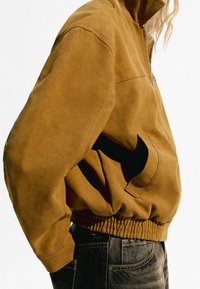 Veste marron à col cheminée avec fermeture éclair, dotée de poches latérales et d'un ourlet élastique. Le matériau semble doux avec une texture lisse.