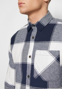 Camisa de franela a cuadros en azul marino y blanco con cuello abotonado, que presenta un bolsillo parche y tela texturizada con patrón de espiga en algunas secciones.