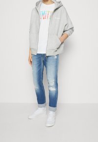 Grå huvtröja över en vit T-shirt med flerfärgad text, kombinerad med ljus tvättade slitna jeans och vita sneakers.