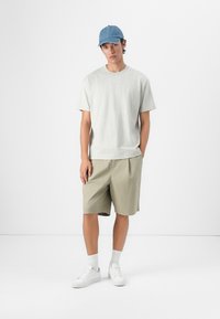 Hellgraues Baumwoll-T-Shirt, beige Shorts, weiße Sneakers und eine blaue Mütze. Das Modell steht vor einem neutralen Hintergrund.