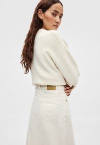 Εκρού φούτερ με χαλαρή εφαρμογή στους ώμους, σε συνδυασμό με λευκή τζιν φούστα. Σημαντικές λεπτομέρειες περιλαμβάνουν μια ετικέτα με λογότυπο στη μέση και υφή με ανάγλυφη όψη.