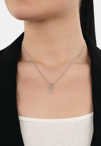 Collana con pendente in argento caratterizzata da un design di spilli di sicurezza intrecciati con piccole gemme trasparenti, su una catena sottile, indossata sopra un top aderente.
