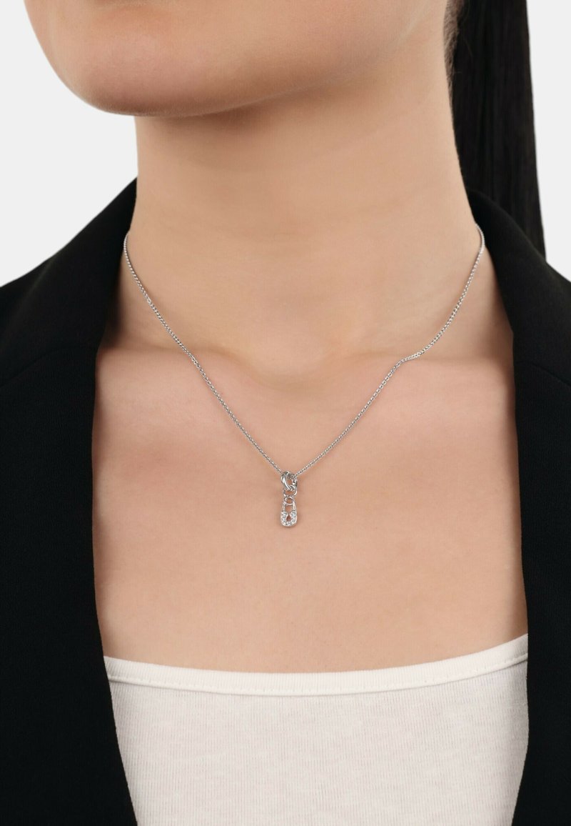 Collana con pendente in argento caratterizzata da un design di spilli di sicurezza intrecciati con piccole gemme trasparenti, su una catena sottile, indossata sopra un top aderente.