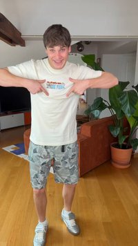 Junger Mann lächelt drinnen, zeigt auf die Grafik auf seinem beigen T-Shirt, trägt Tarnshorts und silberne Turnschuhe, steht auf Holzboden.