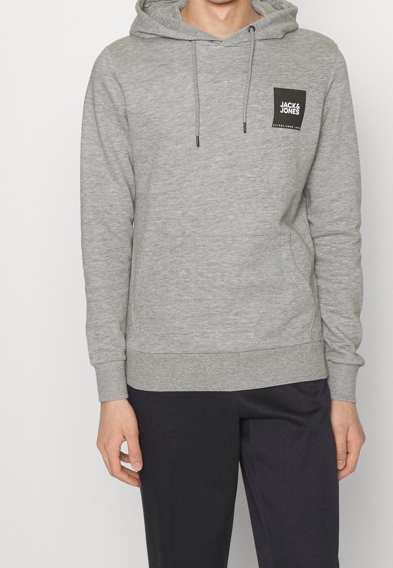 Homme portant un sweat à capuche gris clair avec poche avant et logo noir "Jack & Jones", assorti d'un pantalon noir, debout devant un fond uni.