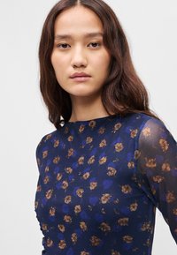 Námořnický top s květinovým vzorem, který obsahuje zlaté a modré květiny. Hladká tkanina s průhlednými dlouhými rukávy a kulatým výstřihem.