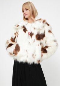 Veste en fausse fourrure avec une base blanche et des taches brunes, dotée d'un col montant, d'une fermeture éclair et de poches latérales. Assortie avec une jupe noire.