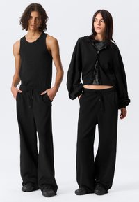 Δύο μοντέλα στέκονται, φορώντας μαύρο παντελόνι με φαρδύ πόδι· το ένα με μαύρο αμάνικο τοπ, το άλλο με κοντό μαύρο hoodie πάνω από ριγέ πουκάμισο.