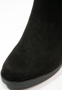 Bottine en daim noir avec une texture lisse, un bout arrondi et une semelle en caoutchouc basse. Aucune ferrure ni motif visible. Vue en gros plan.