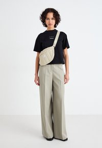 Svart kortärmad skjorta med vit text, beige kviltad axelväska, ljus khaki vida byxor och neutral bakgrund.