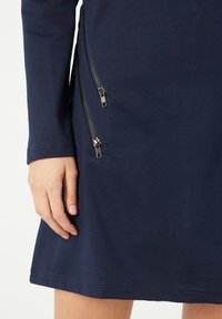 Vestido azul marino con mangas largas y corte recto. Presenta dos bolsillos laterales con cremallera y detalles de metal. Textura de algodón suave.