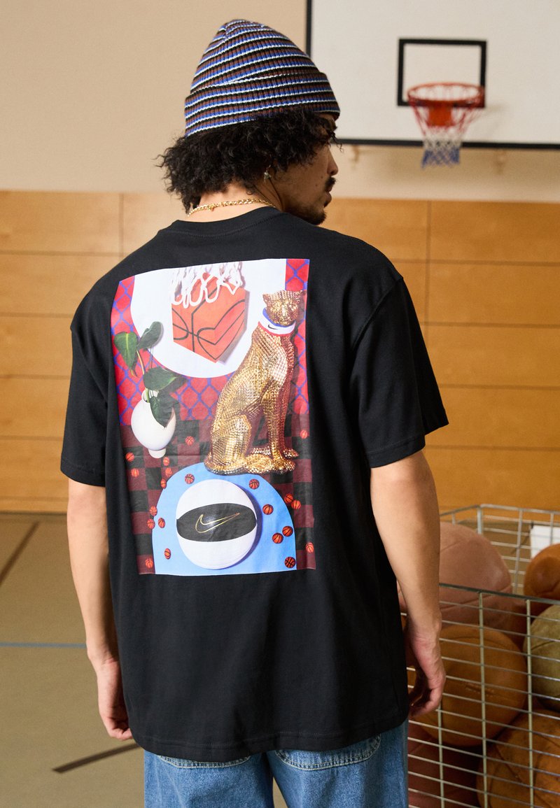 Camisa negra de manga corta con un gráfico colorido que presenta un gato dorado, balones de baloncesto y el logo de Nike sobre un fondo con patrón.