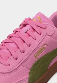 Artimas rožinių zomšinių sportbačių su žalia juosta, rožinėmis raištelėmis ir aukso spalvos Puma logotipu pusėje ir liežuvėlyje, baltame fone.