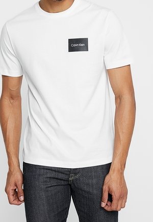 White short-sleeve t-shirt made of cotton, featuring a black rectangular logo patch with white text on the left chest area.
