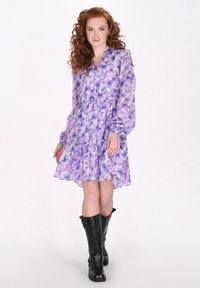 Leichtes, luftiges Kleid mit lila Blumenmuster, langen Ärmeln, V-Ausschnitt und gestuften Saum. Trägt man mit kniehohen schwarzen Stiefeln.
