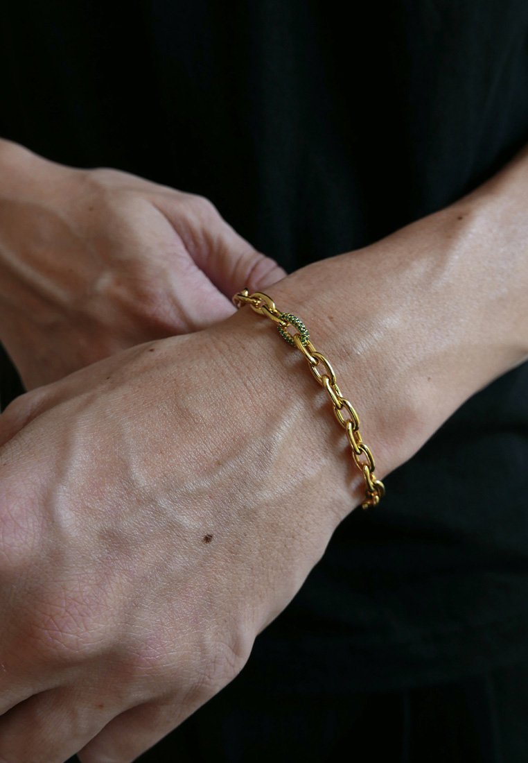 Χρυσή αλυσίδα βραχιόλι με οβάλ συνδέσμους, που διαθέτει μία μικρή πράσινη λεπτομέρεια. Φορεμένο σε καρπό με ορατή υφή δέρματος.