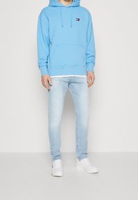 Sudadera azul clara con bolsillo delantero y cordón, combinada con jeans de lavado claro y zapatillas blancas. La sudadera presenta un pequeño logo.