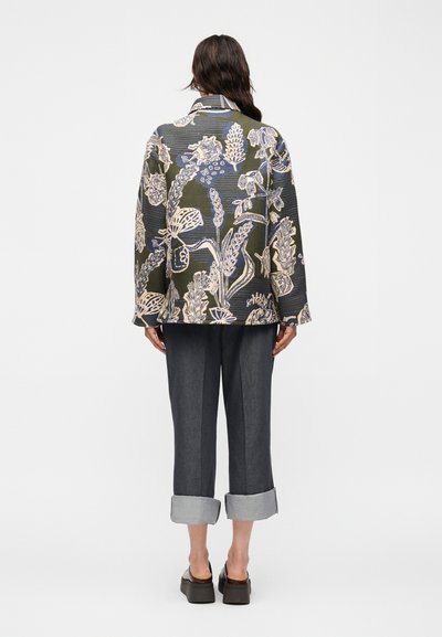 Γυναίκα που φορά μακριμάνικο σακάκι με λουλουδάτο σχέδιο, σκουρόχρωμο κατεβασμένο παντελόνι έως τον αστράγαλο και μαύρα παπούτσια πλατφόρμα, στέκεται γυρισμένη με την πλάτη σε λευκό φόντο.