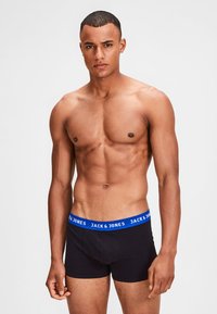 Schwarze Baumwoll-Boxershorts mit einem blauen elastischen Taillenbund, auf dem "JACK & JONES" in Weiß steht. Das Modell steht selbstbewusst da und zeigt eine durchtrainierte Figur.