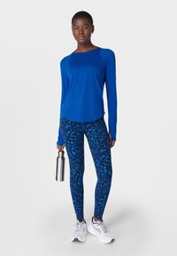 Blauw langmouwtop en blauwe gestreepte leggings, met een gladde textuur. Model houdt een zilveren waterfles vast en draagt witte sneakers.