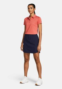 Sarkans heather polo krekls ar apkakli, apvienots ar tumši zilu svārku. Modelis nēsā baltas sporta kurpes. Vienkāršs, funkcionāls dizains.