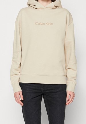 Personne portant un sweat à capuche beige Calvin Klein avec la capuche baissée et un pantalon noir, debout devant un fond clair uni.