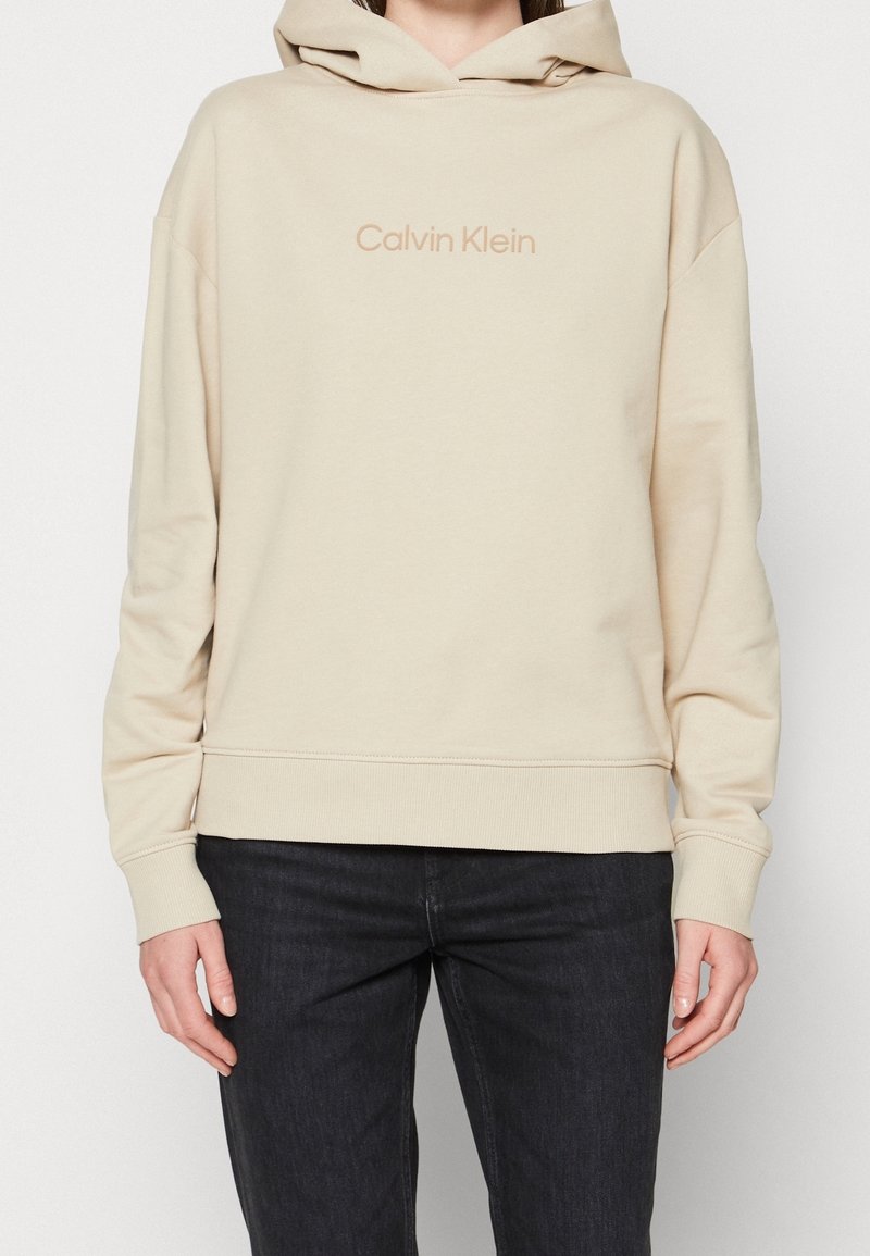 Personne portant un sweat à capuche beige Calvin Klein avec la capuche baissée et un pantalon noir, debout devant un fond clair uni.