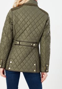 Gequiltede olijfgroene jas met hoge kraag, ceintuurlijn, gouden hardware accenten en een diagonale stiksels patroon op de stof.