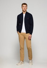 Marinfärgad zip-up-tröja över en vit skjorta, parat med khakifärgade byxor och ljusa sneakers. Notabel detalj: liten logotyp på tröjan.