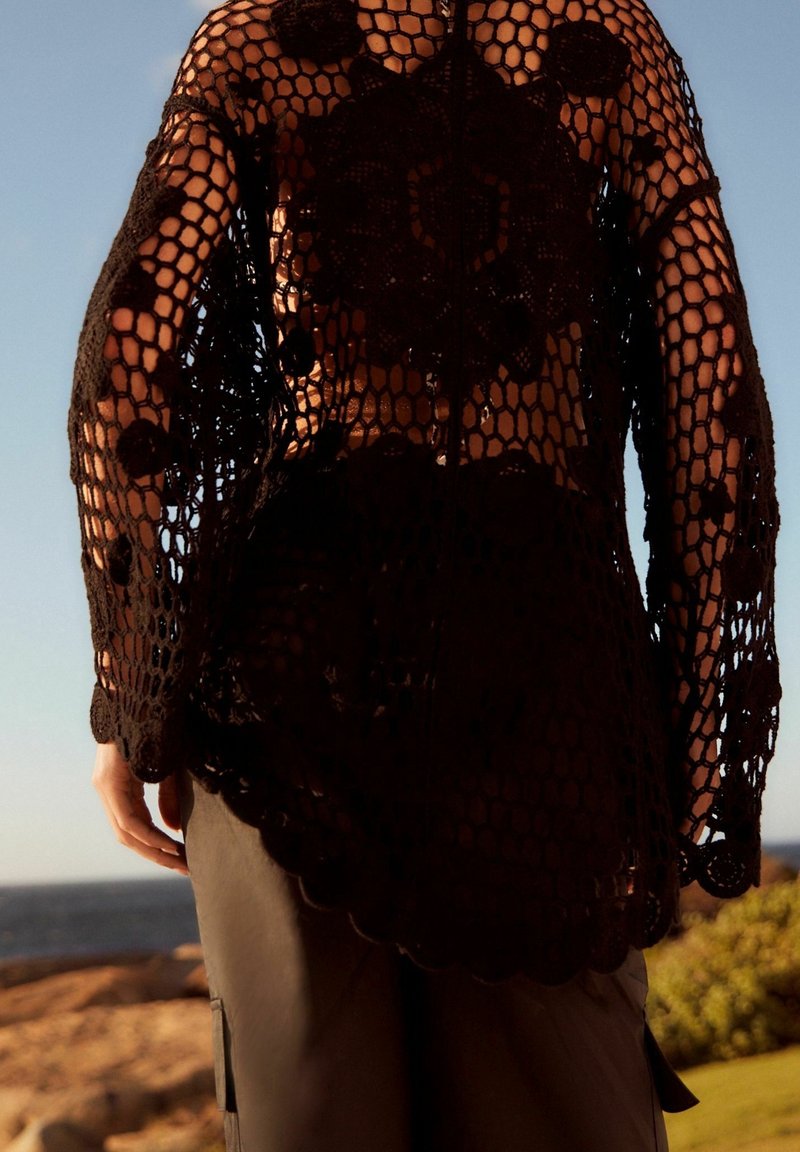 Czarny top z szydełka o otwartym, plecionym wzorze; falbaniasty dół i długie rękawy; w połączeniu z ciemnymi spodniami; tło przedstawia naturalny krajobraz.